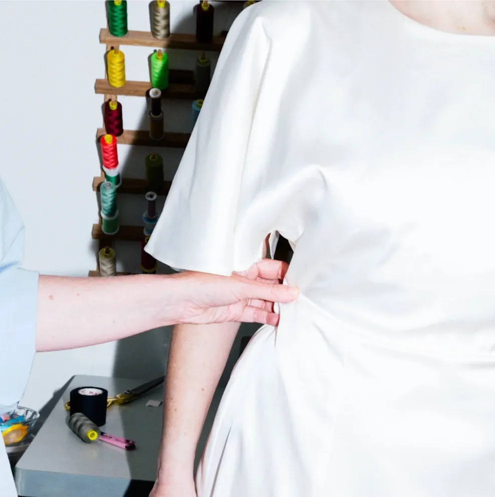 A hand easing the side seam of a white dress, with a rack of coloured thread spools behind