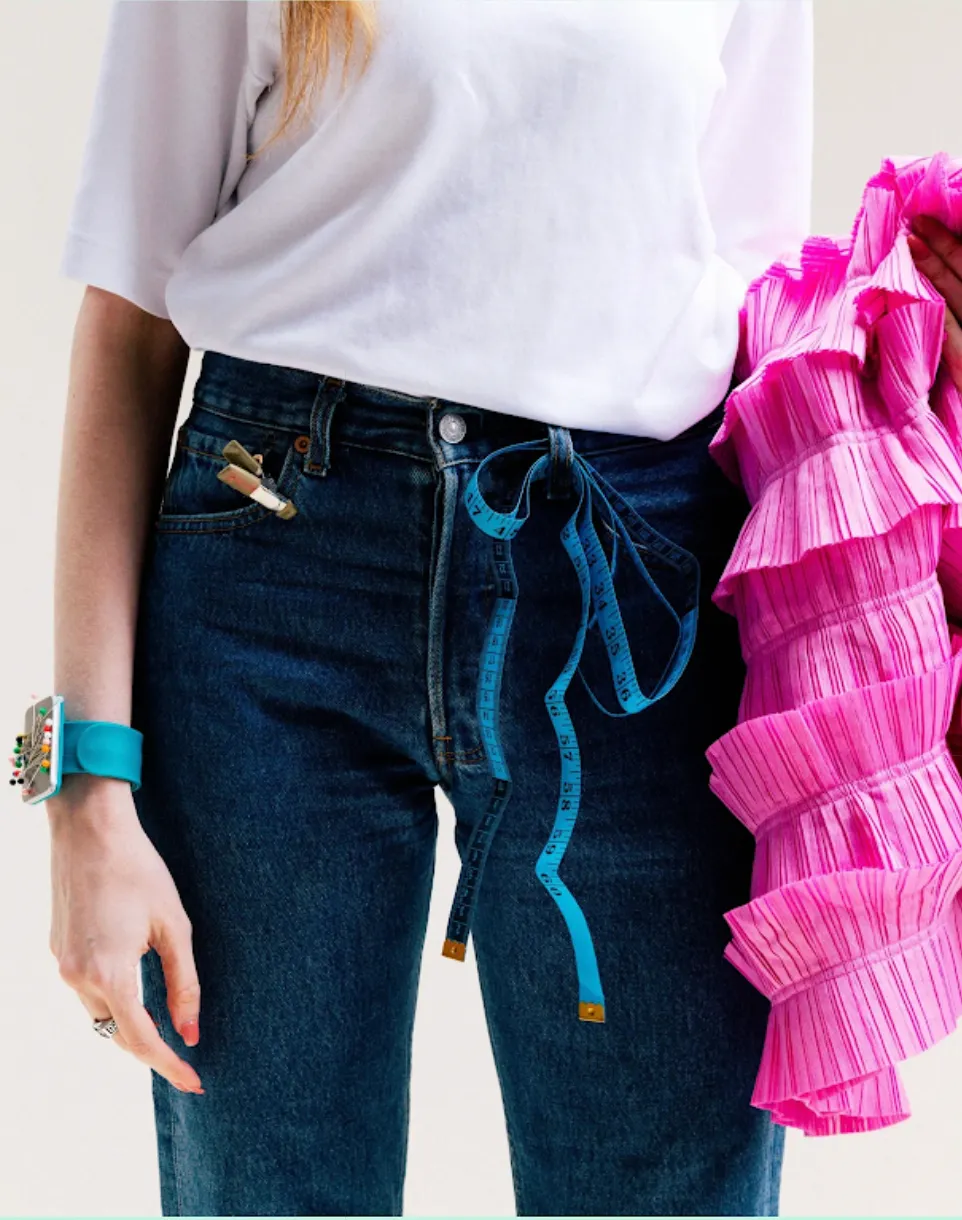 A tailor in jeans with a tape measure at the waist and a pin cushion on the wrist, holding a pink pleated dress over one arm